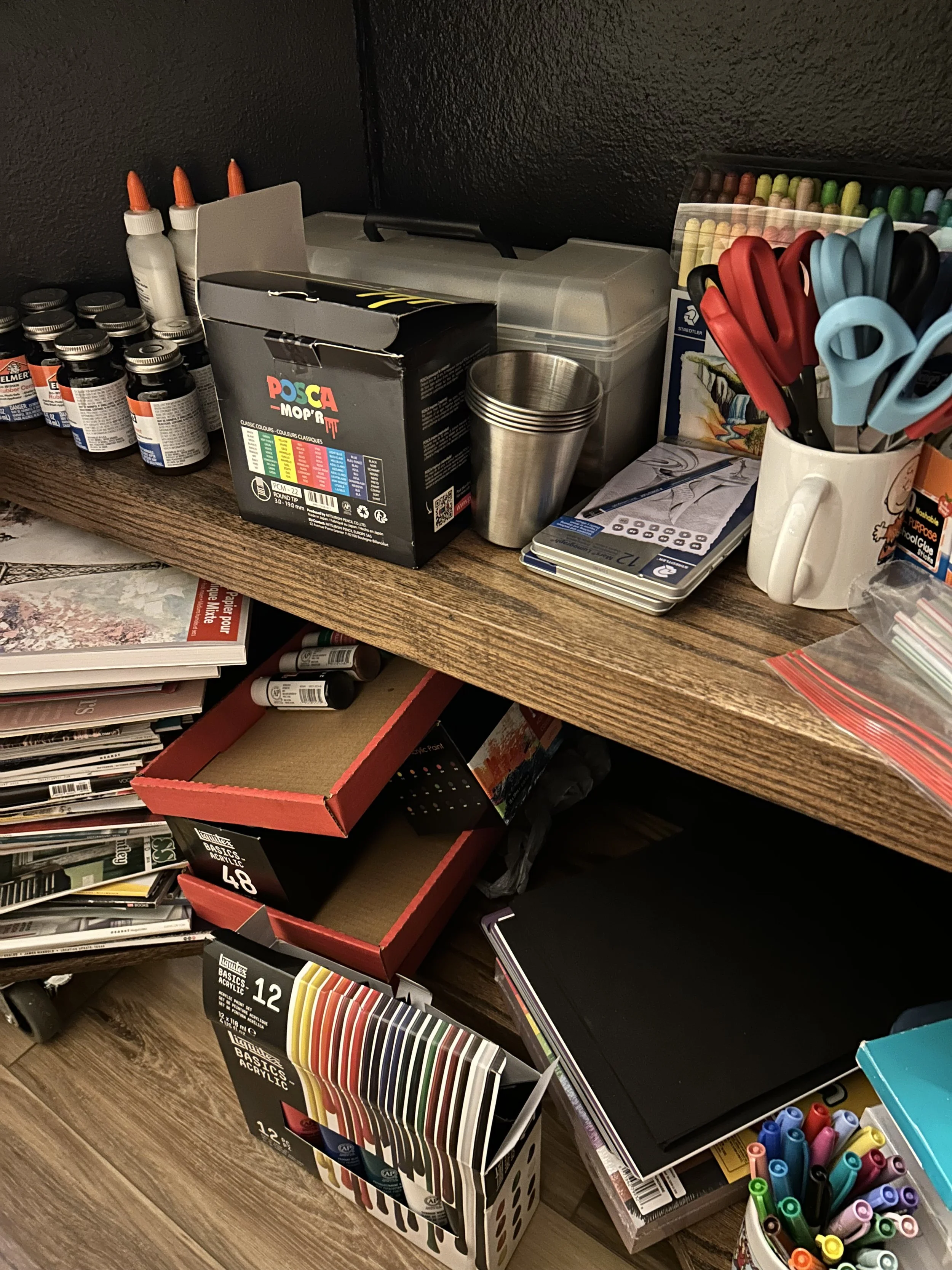 Craft and art supplies on a wooden desk, including paint bottles, glue, scissors, markers, coloring books, and storage containers.
