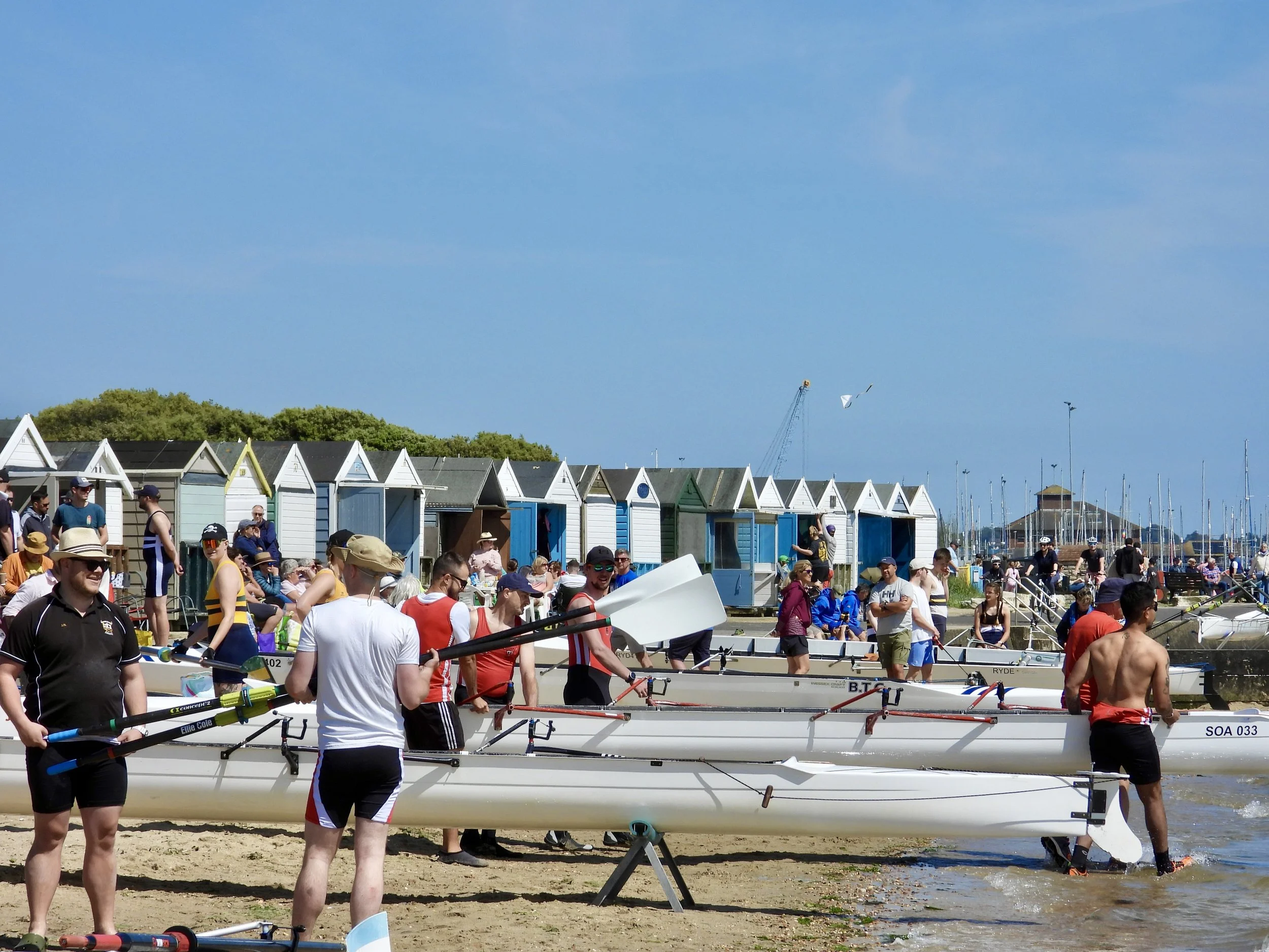 People preparing rowing boats on a beach with beach huts and marina in the background on a sunny day.