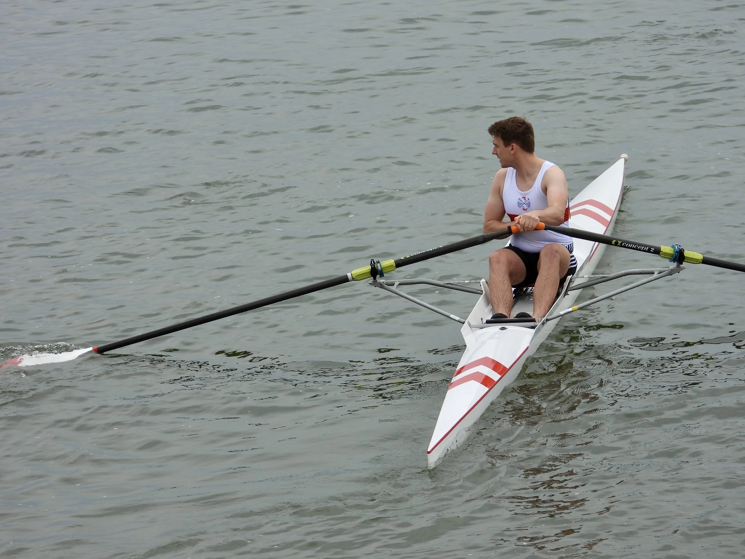 A man in a white sleeveless shirt and black shorts sailing a racing shell on a body of water.