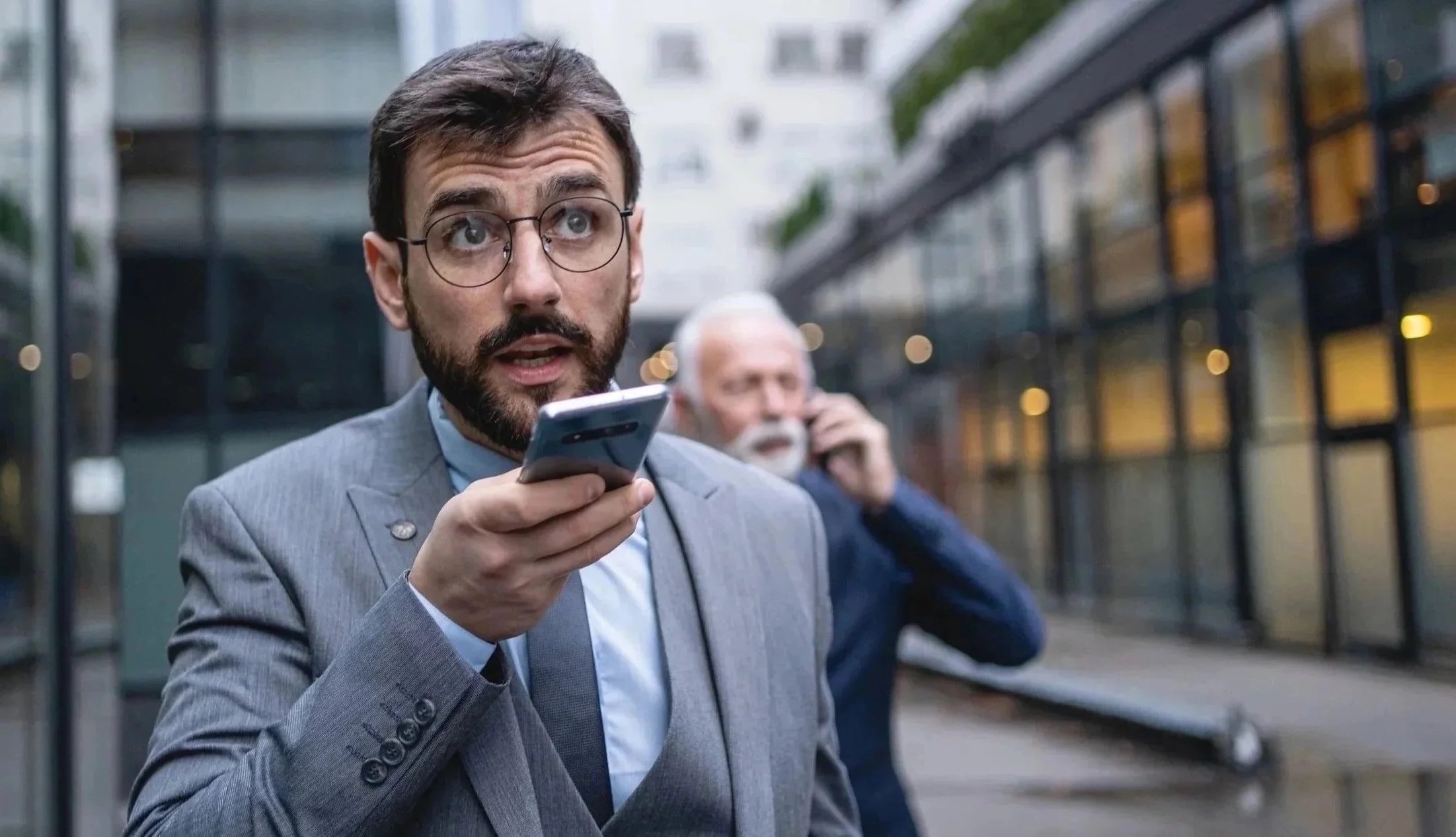 Yupa® Coach A man in a gray suit and glasses holding a smartphone, looking surprised, with another man in the background on a city street.