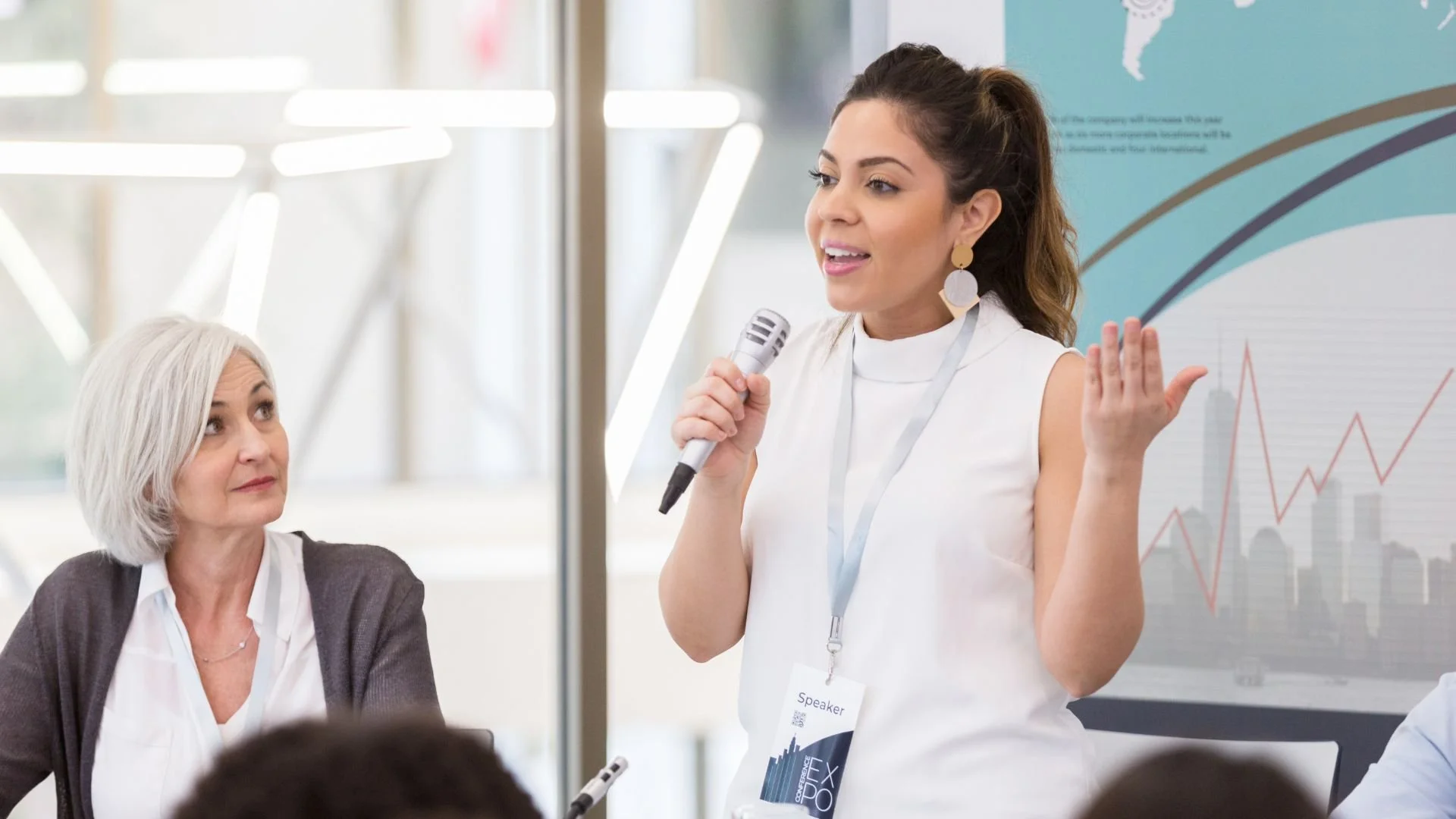Yupa® Coach A young woman speaker holding a microphone and gesturing with her hand while addressing an audience at a conference, with an older woman seated nearby and a presentation slide with graphs in the background.