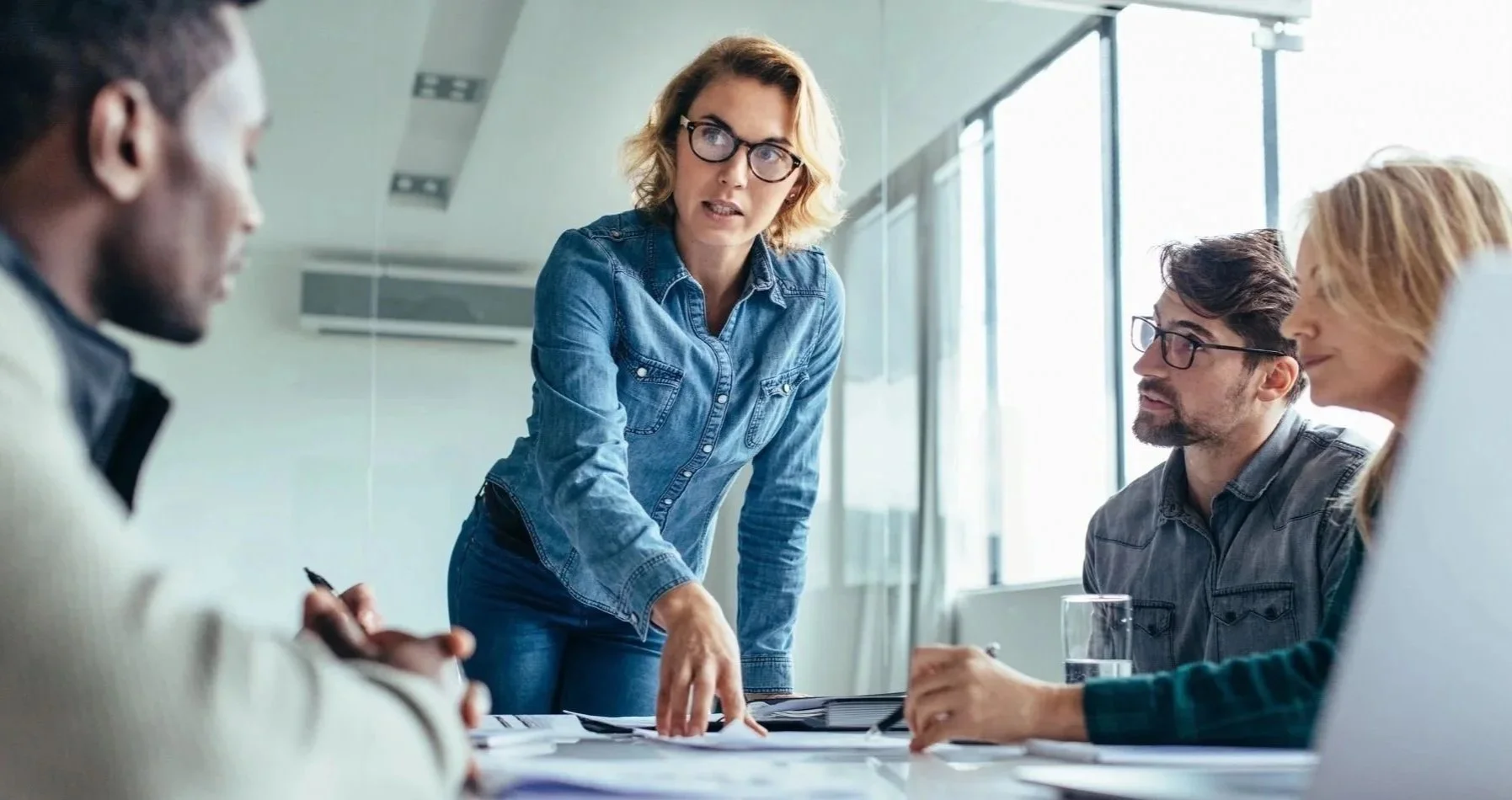 Yupa® Coach A woman is standing and talking to a group of three people sitting around a table during a meeting in an office.