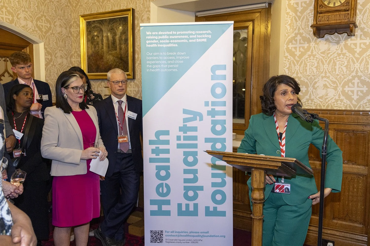 Woman in green suit speaking at a podium during a conference, with a banner behind her that reads "Health Equity Foundation." Several people are listening, some holding glasses of wine, in a wood-paneled room with wallpaper and artwork.