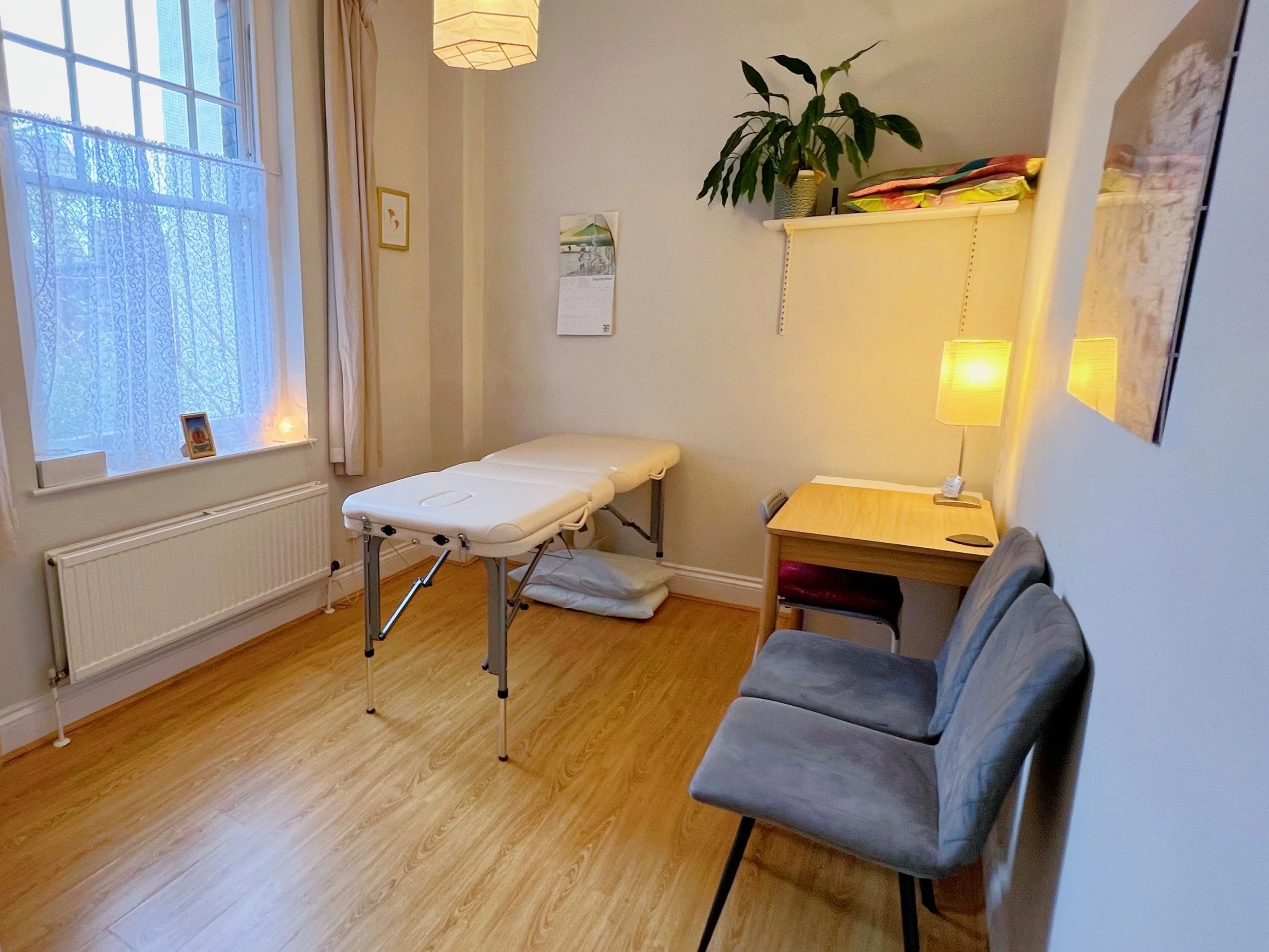 A cozy practice room called "Room 2" with a massage couch, two chairs, a wooden desk with a table lamp, a potted plant, and a window with sheer curtains.