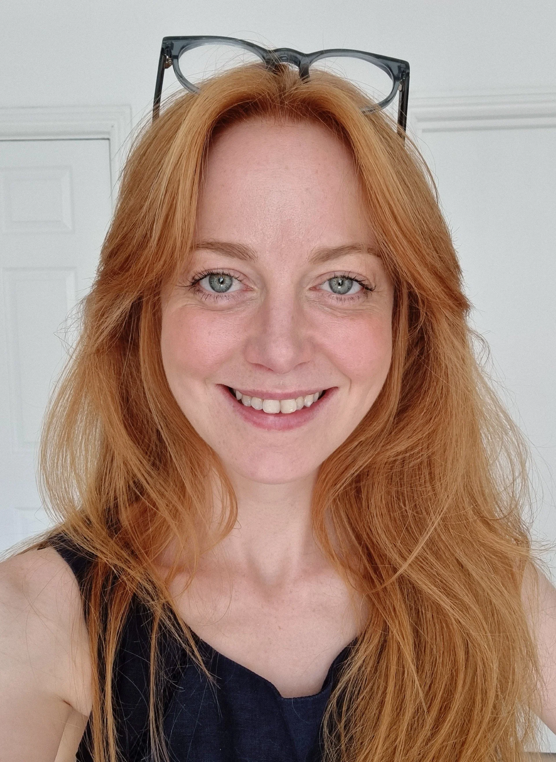 Morag Ruffell, a woman with long red hair, glasses, and a gold necklace smiling at the camera. She is wearing a light-colored top. There are green leaves and fairy lights in the background.