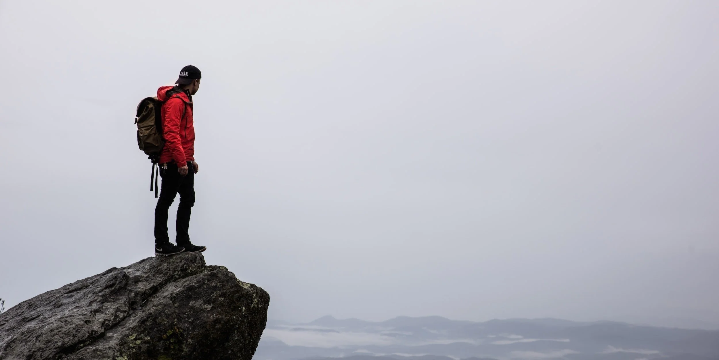 A person standing on the edge of a rocky cliff, looking into the cloudy sky, with mountains and fog in the distant background.