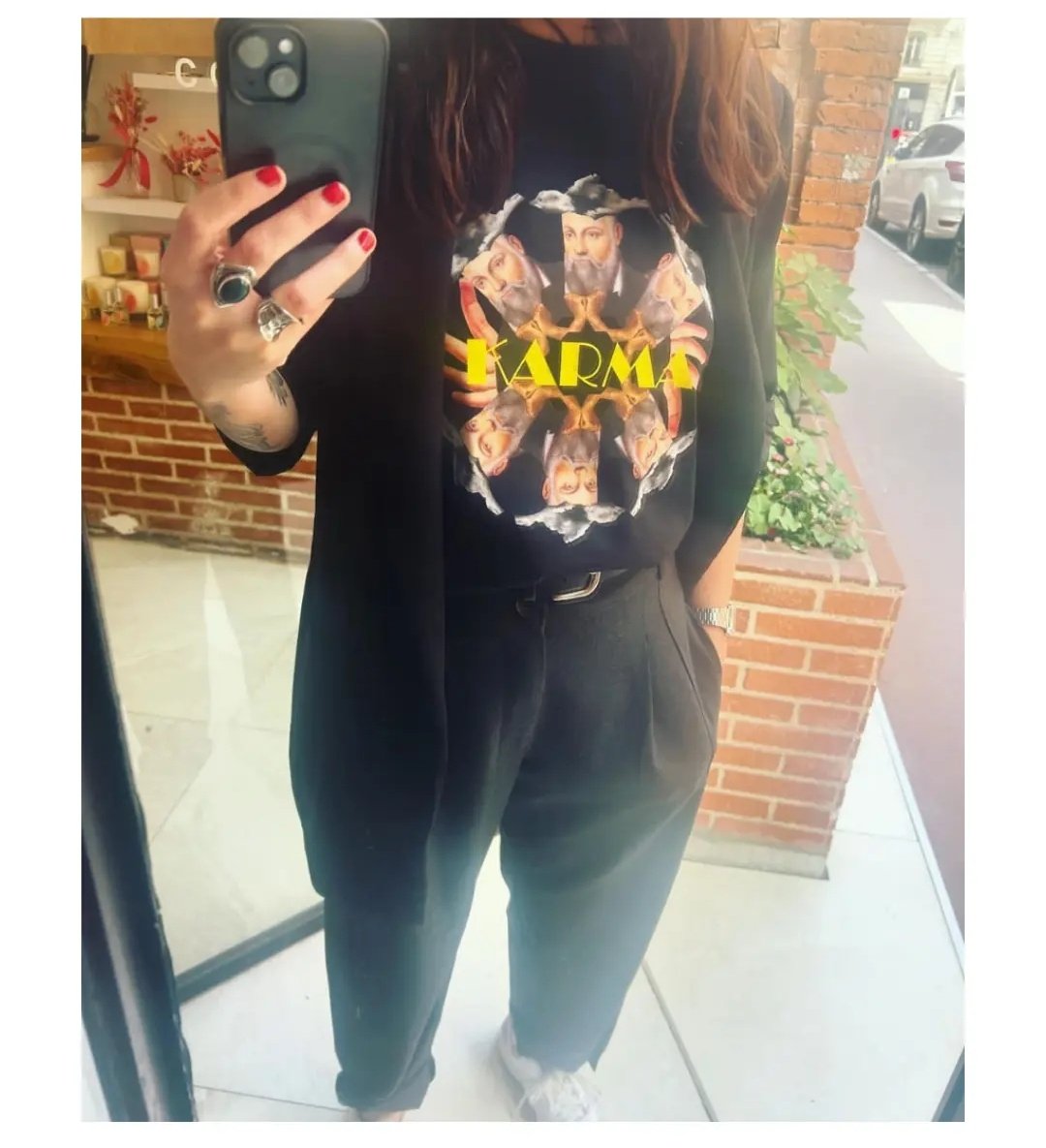 Une femme prenant une photo d'elle-même dans un miroir. Elle porte un t-shirt noir avec un design coloré de visages et la mention "karma" en jaune. Elle a des cheveux bruns, porte des bijoux et a les ongles rouge. Le fond montre une boutique avec des décorations et des voitures à l'extérieur.