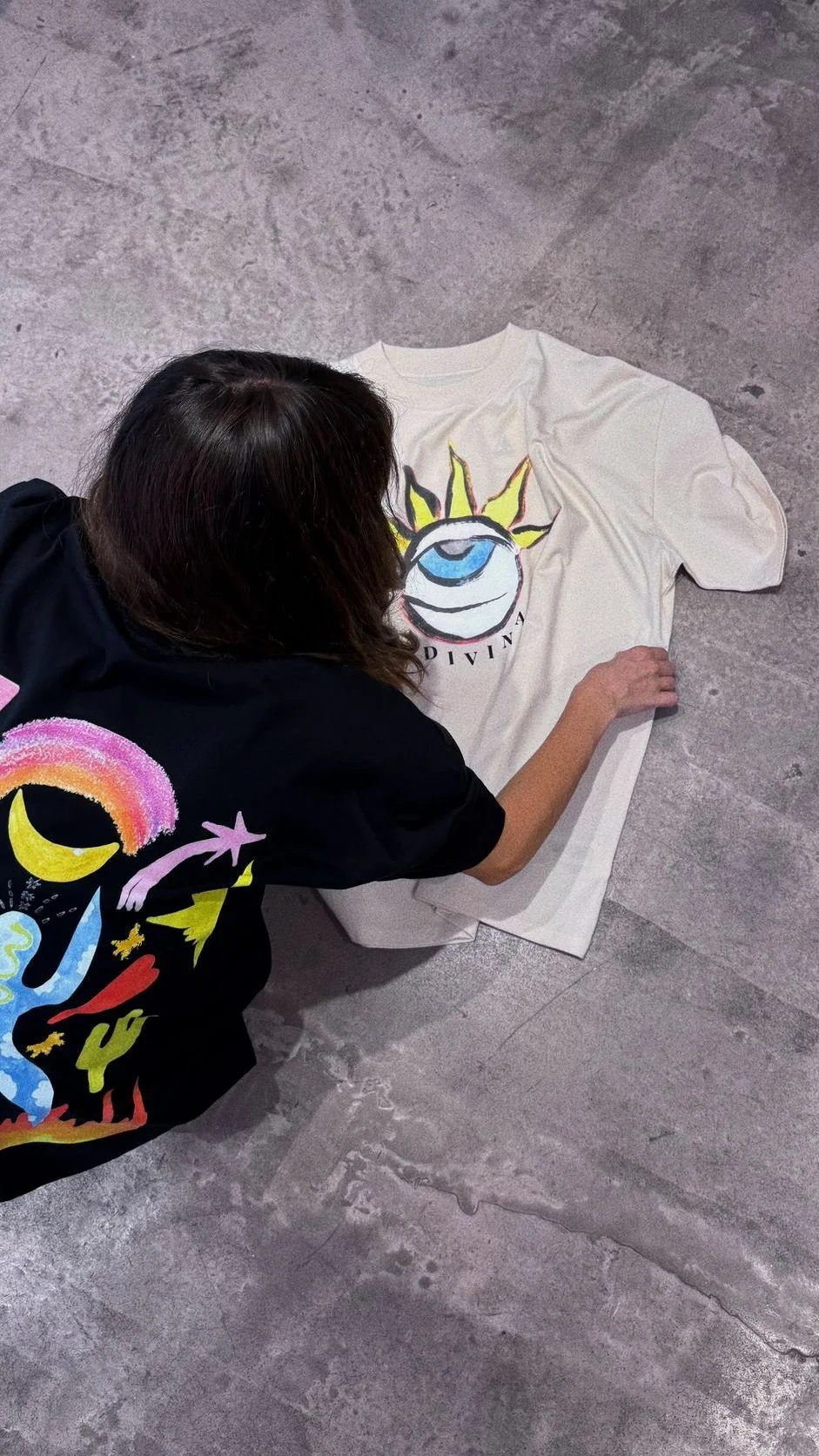 Une femme dessine ou peint un design coloré sur un t-shirt blanc posé sur le sol en carrelage gris.