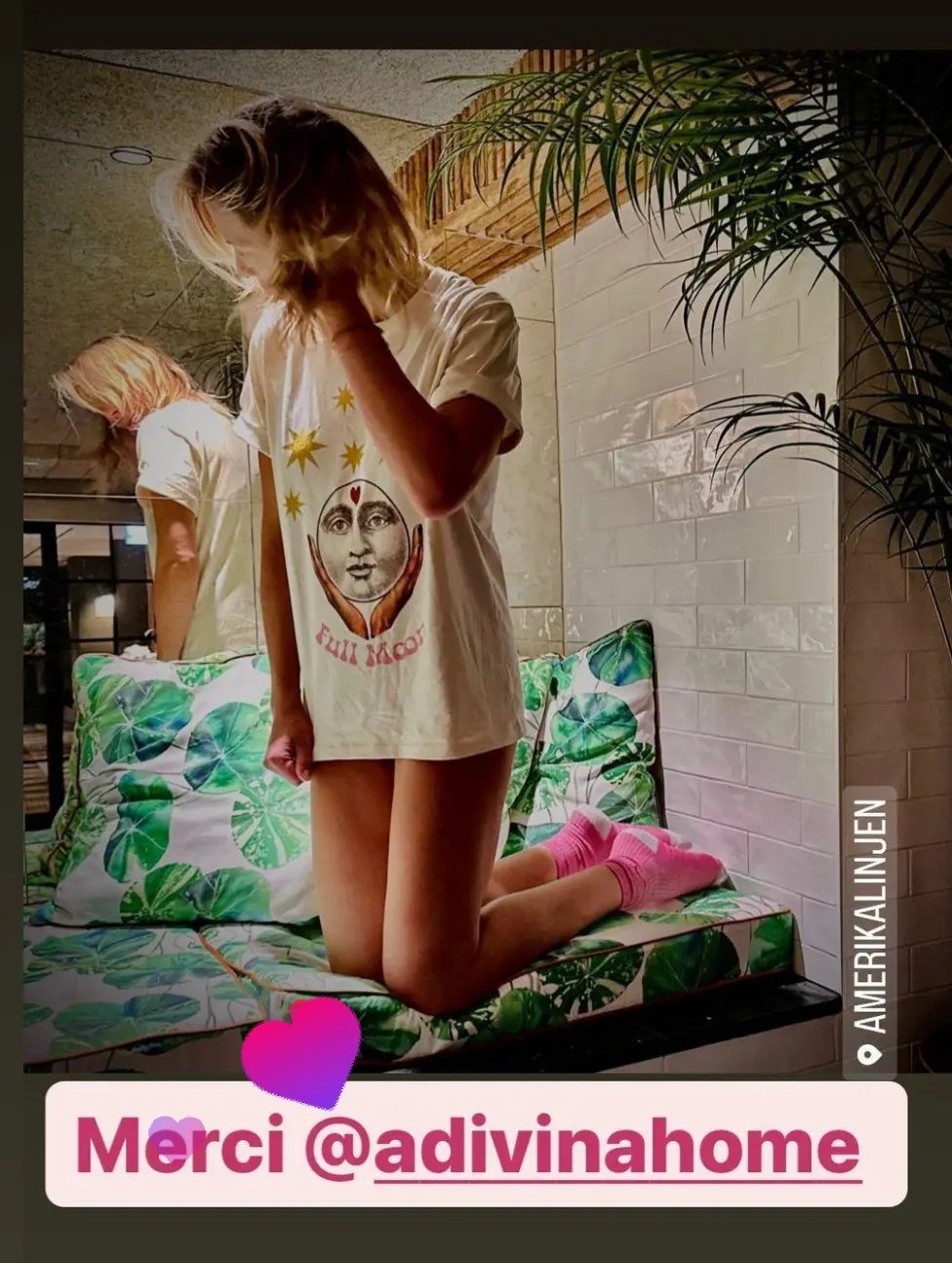 Deux jeunes femmes portant des t-shirts assortis avec des motifs artistiques, l'une à genoux sur un canapé avec des coussins à motifs de feuilles vertes, dans un intérieur chaleureux avec des plantes et une paroi en briques blanches.