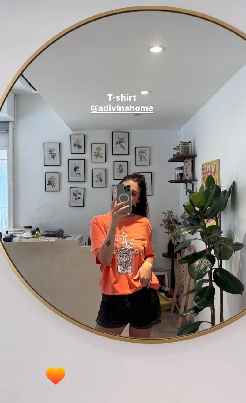 Une femme prend un selfie dans un miroir rond. Elle porte un t-shirt orange avec un dessin d'éléphant et des shorts noirs. La pièce a un mur avec plusieurs cadres photo de fleurs, une plante verte à droite, et des étagères avec des objets décoratifs.