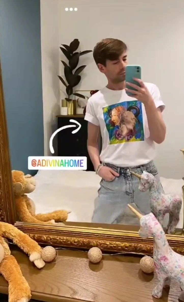 Un jeune homme se prend en selfie dans un miroir, portant un t-shirt avec une impression artistique et tenant une main dans la poche. Sur la commode en dessous, il y a deux girafes en peluche, un chat en peluche et plusieurs boules de laine. À l'arrière, un meuble avec une plante verte en pot.