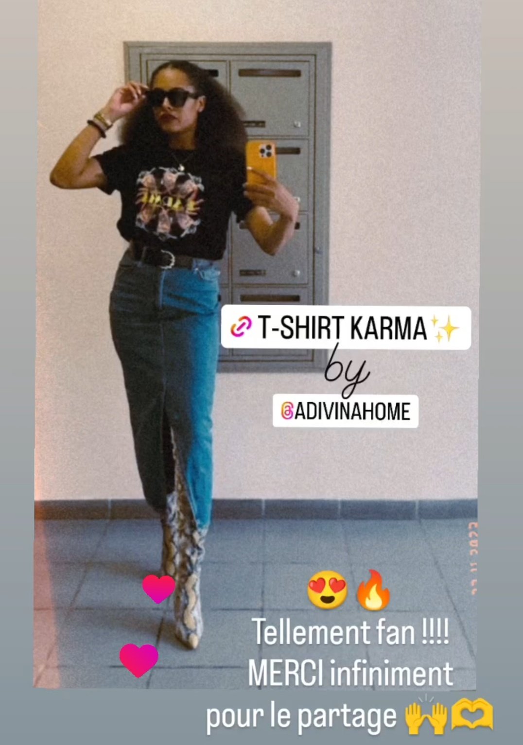 Une jeune femme pose devant un miroir, portant un t-shirt noir avec un design floral et un pantalon en denim avec des bottes à imprimé serpent. Elle porte des lunettes de soleil et tient un téléphone jaune pour prendre la photo.