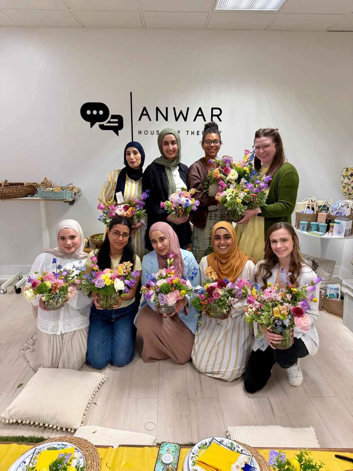 Group of nine women holding bouquets of colorful flowers in a room with a sign that reads 'ANWAR House of the' on the wall behind them.