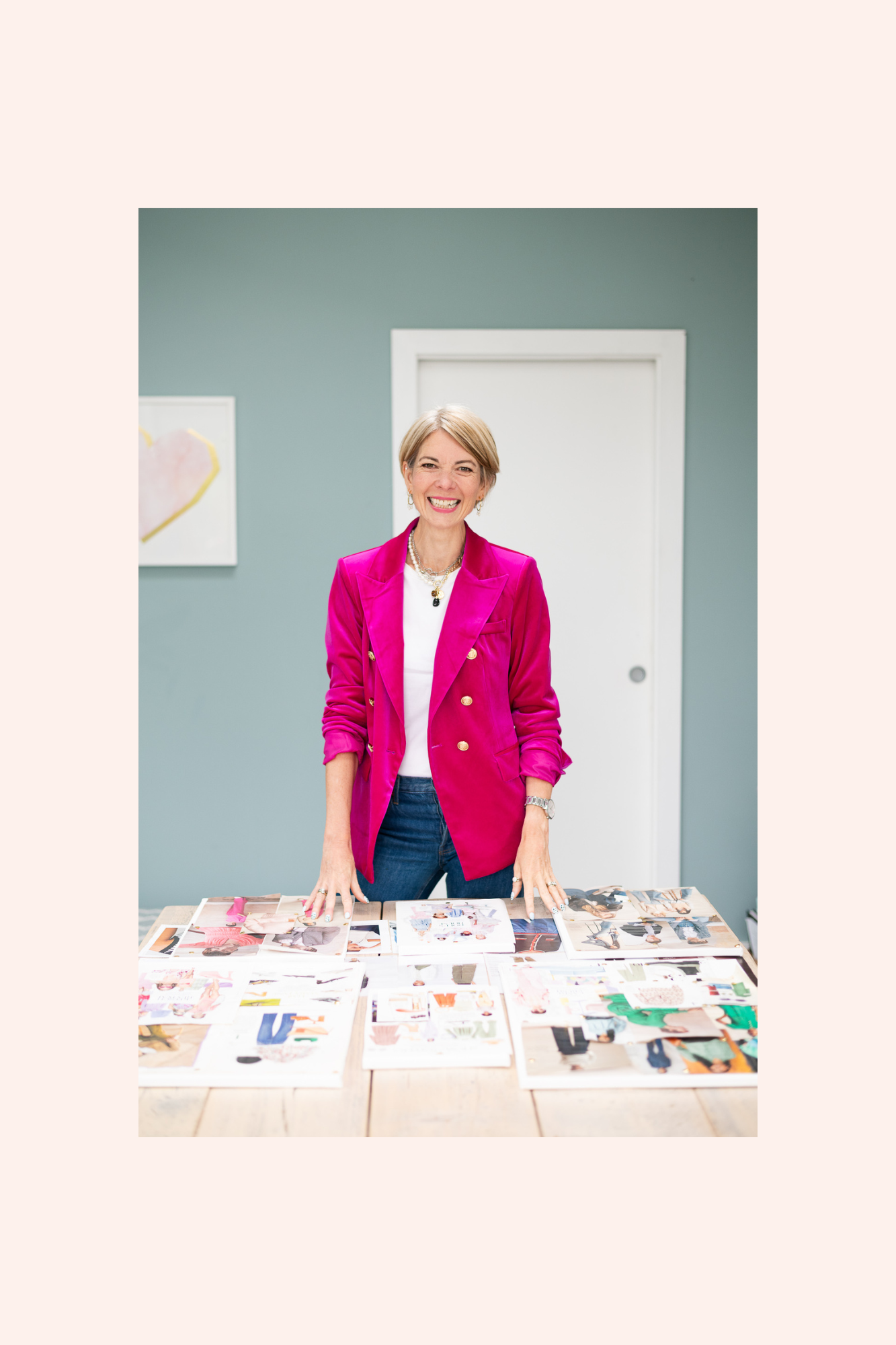 A woman with short blonde hair, wearing a bright pink blazer over a white top, standing behind a table with fashion magazine images spread out, smiling in a room with a light green wall and a white door.