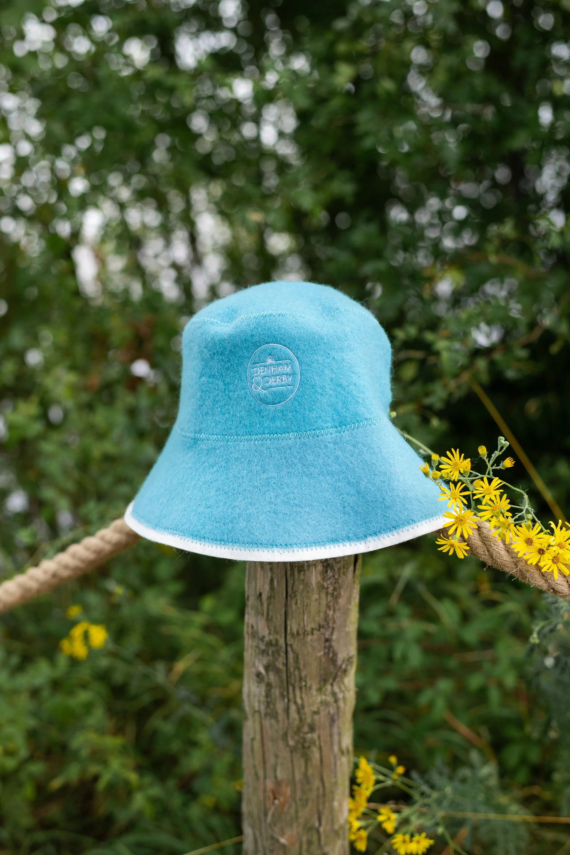 A light blue bucket hat with the words 'DENTHAM DERBY' embroidered on it, placed on a wooden post with yellow flowers nearby, outdoor setting with green foliage in the background.