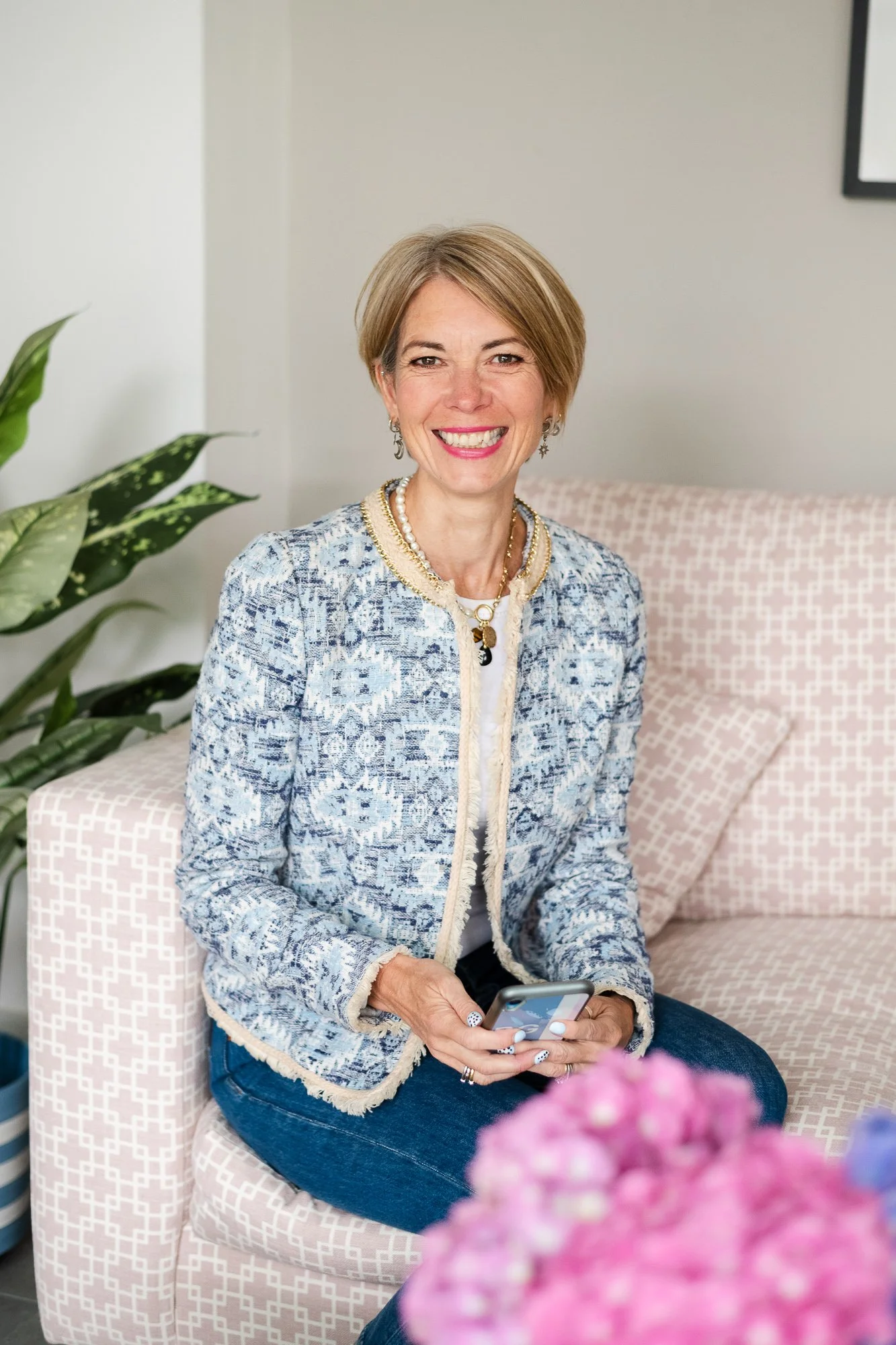A smiling woman with short blonde hair sitting on a pink patterned sofa, holding a smartphone, in a room with a large green plant and pink flowers in the foreground.