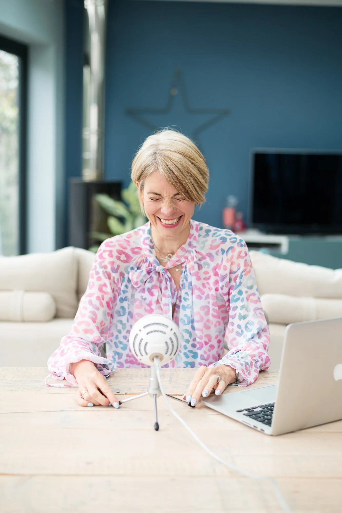 A woman with short blonde hair smiling and laughing while sitting at a wooden table with a microphone and a laptop in front of her. She is wearing a colorful leopard print blouse and appears to be recording a podcast or participating in an online video call, in a bright, modern living room with a blue wall and TV in the background.