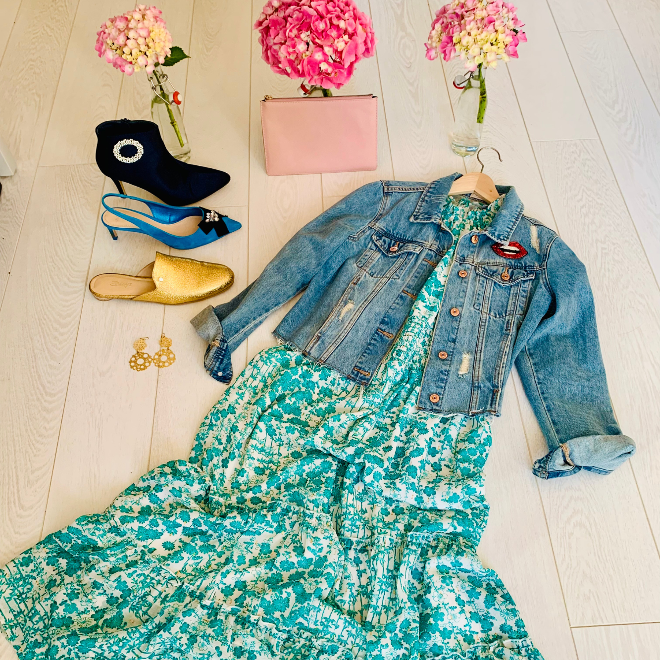 A dress with a denim jacket on top laying next to shoes, earrings, a purse, and flowers