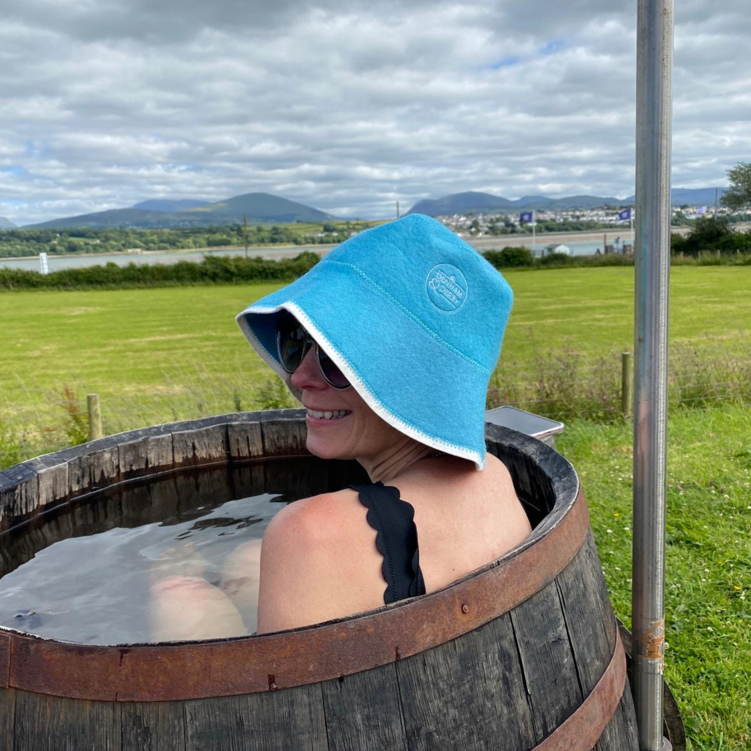 Woman sitting in a wooden tub filled with water outdoors, wearing sunglasses, a black swimsuit, and a blue towel hat, with a scenic landscape of green fields, water, hills, and a cloudy sky in the background.