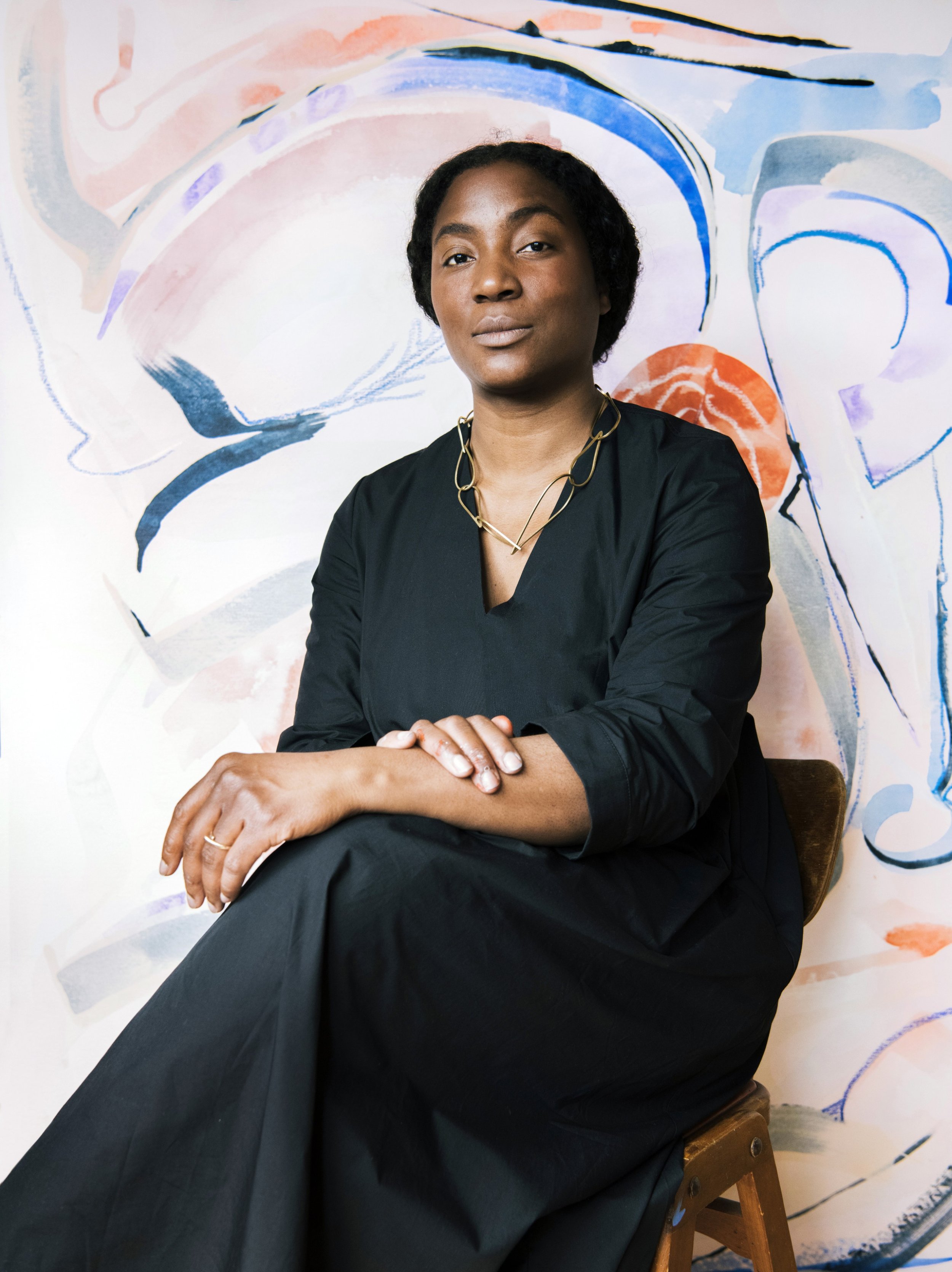 Artist, Simone Brewster, is sat in front of one of her large abstract paintings. The abstract painting features soft, colourful gestural brushtrokes representing abstracted female forms and internal psychological and emotional landscapes