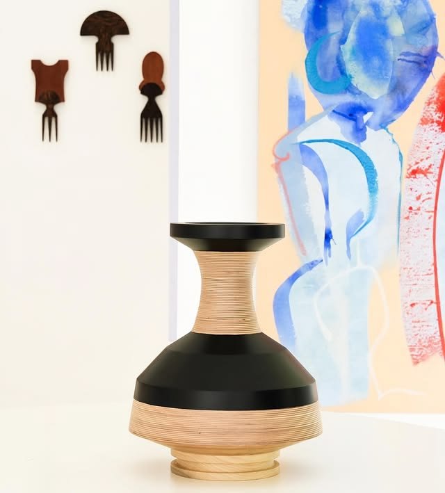 A decorative ceramic vase with a black top and beige ribbed middle section, placed on a white surface. In the background, a colorful abstract painting and four small wall-mounted wooden fork-shaped sculptures are visible.