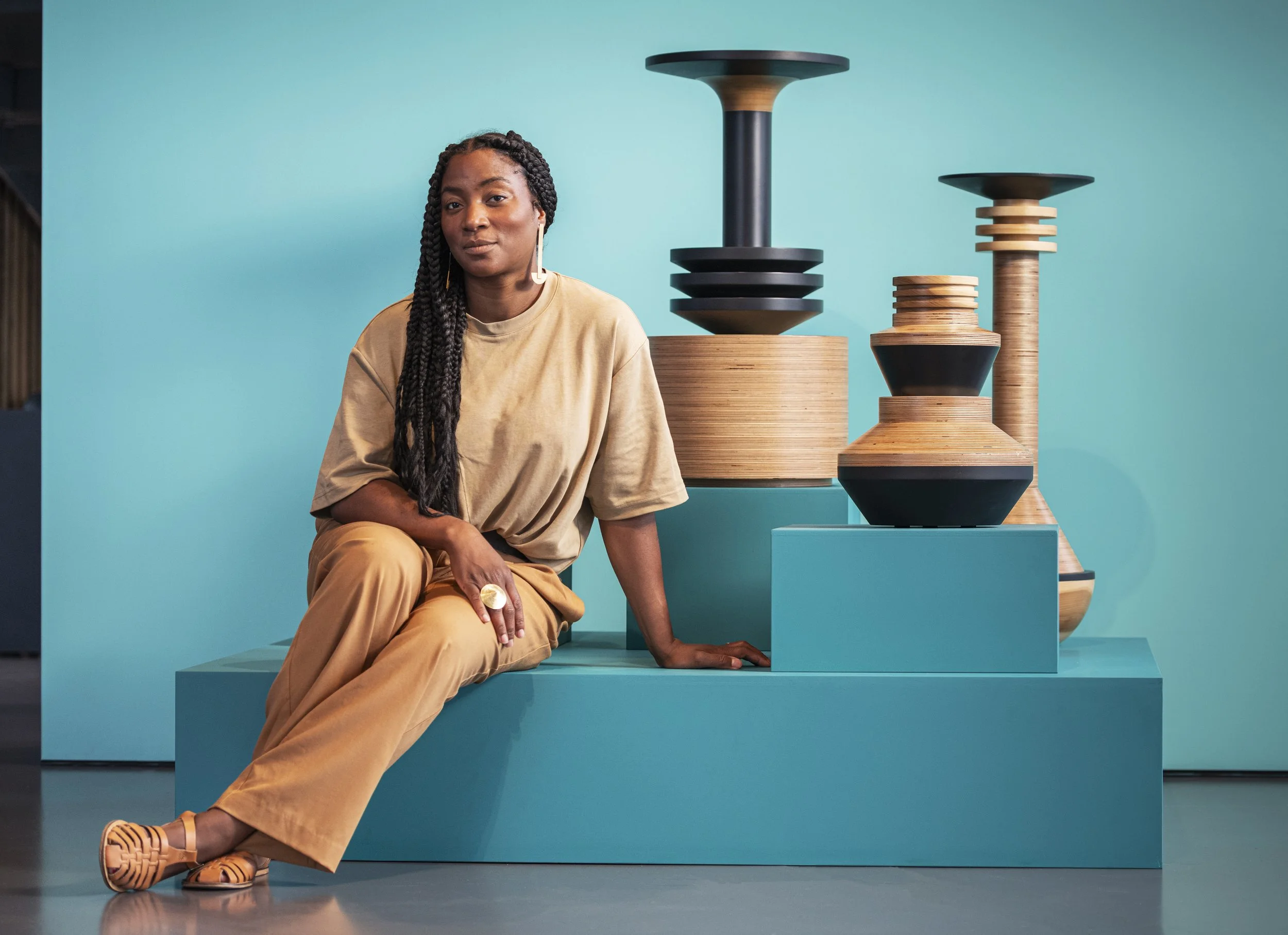 A woman with long braided hair, wearing a beige t-shirt and tan pants, sits on a turquoise platform in front of a turquoise background. She is posed with her left arm resting on the platform and her right arm on her knee. To her right are modern, geometric wooden and black objects with various shapes and sizes, displayed on turquoise stands.