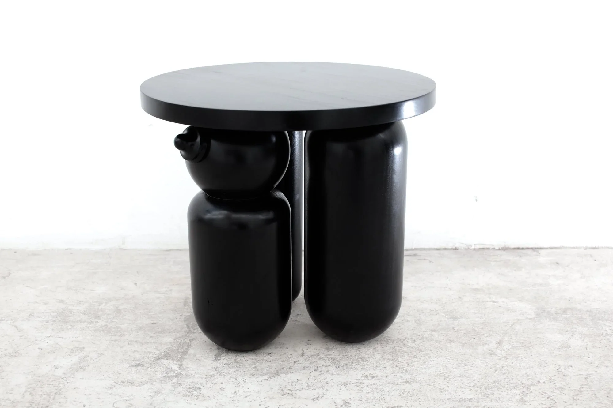 A modern black side table with a round white marble top and three sculptural black bases. The table is placed on a concrete floor against a plain white wall.
