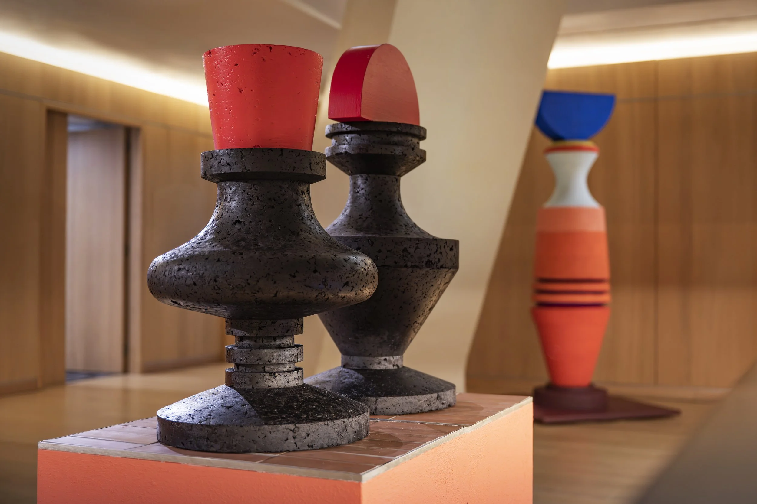 Three modern sculptures in a gallery, made of textured materials with geometric shapes and bold colors, arranged on a wooden platform.
