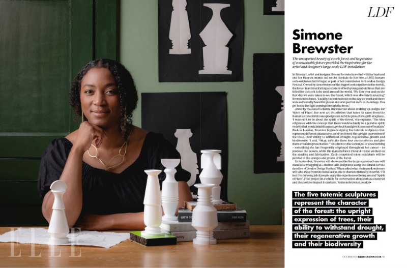 Simone Brewster, sitting at a table with white sculptural objects and books, in front of a green wall with black and white art pieces, next to a feature article in Elle magazine. Features small totemic sculptures designed for public art installation.