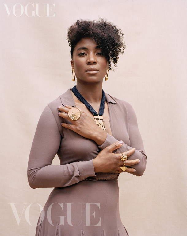 Artist Simone Brewster is wearing gold jewelry, including earrings, a large ring, and a necklace (designed by the artist), poses for a Vogue photoshoot against a light pink background. Feature for Black British Jewellery Design in Vogue.