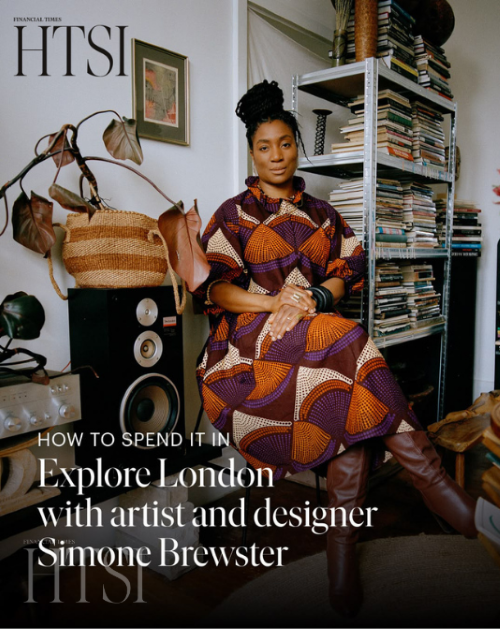Artist Simone Brewster sitting in a room with a large bookshelf filled with books, wearing a colorful patterned dress and brown boots, with a speaker and plants nearby. Artist featured in Financial Time's How To Spend It.