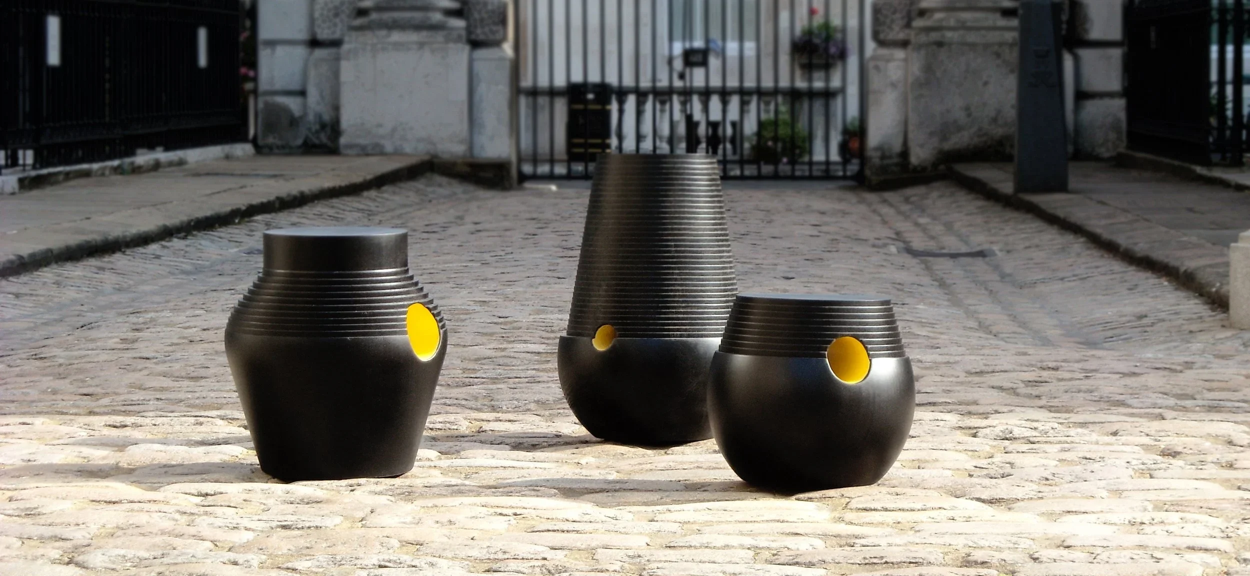 Three step stools with yellow holes in a cobblestone courtyard designed by artist Simone Brewster, with an iron gate and stone pillars in the background.