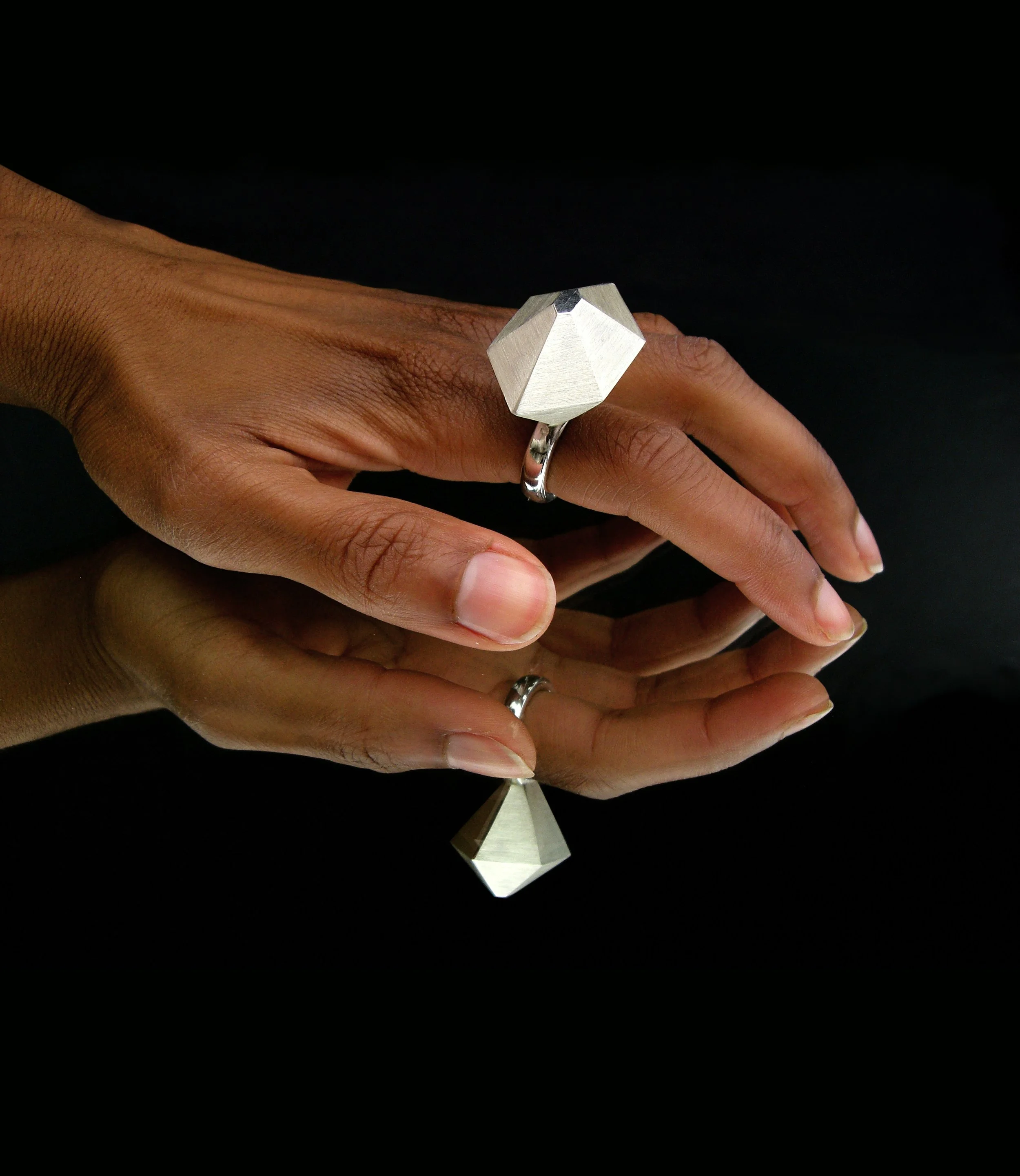 Close-up of a hand wearing a silver geometric ring with a large, metallic, triangular-shaped form against a black background.