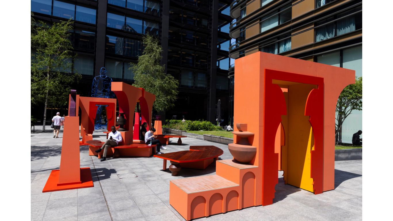 Large scale public installation by Simone Brewster. Orange coloured architectural structures built with cutouts silhouettes of sculptural vessels and archways stand amidst seating area that has also been built into the installation. 