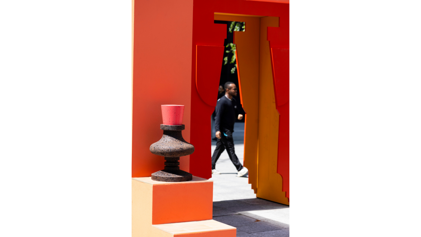 In the foreground there is a cork sculpture that rests on area of the public installation that features tiled steps. The backdrop to the sculpture is a large orange architectural form that has a tall cutout archway in the shape of a vessel