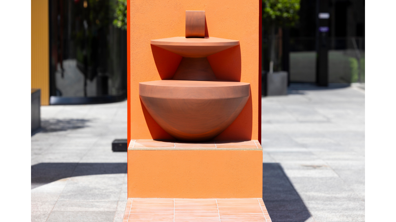 A straight on image of a scuplture made of wood that rests of a tiled step of the public installation. A close up part of the public art installation has been captured with the local environment visible in the background.