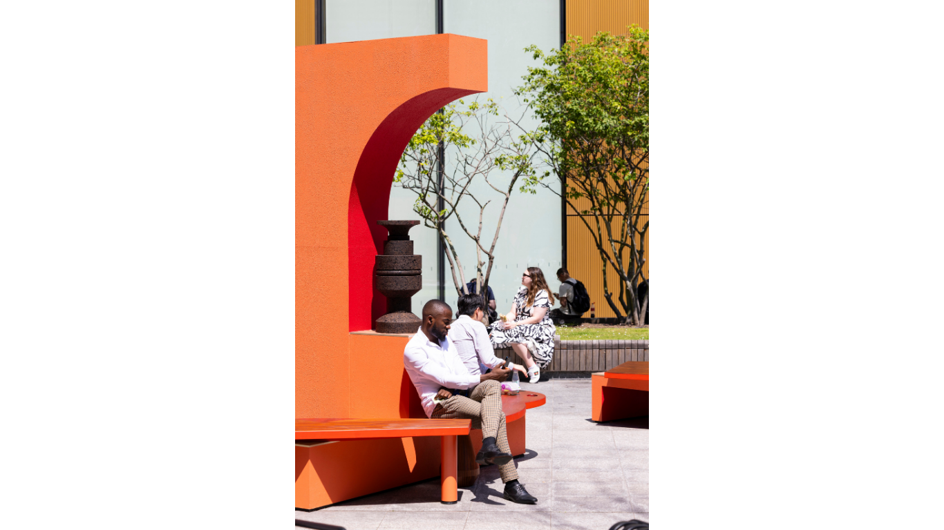 A public art installation in London by artist Simone Brewster. The image sees the seating area of the public installation with a couple people seated on the bench part of the structure. Behind one man's head is a cork sculpture.