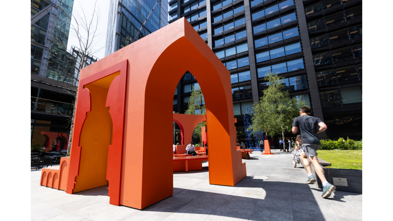 A public art intallation in London by artist Simone Brewster. A large part of the orange architectural structure is in the foreground. Through one archway you see people are seated on benches that are a part of the installation. A man is running by.