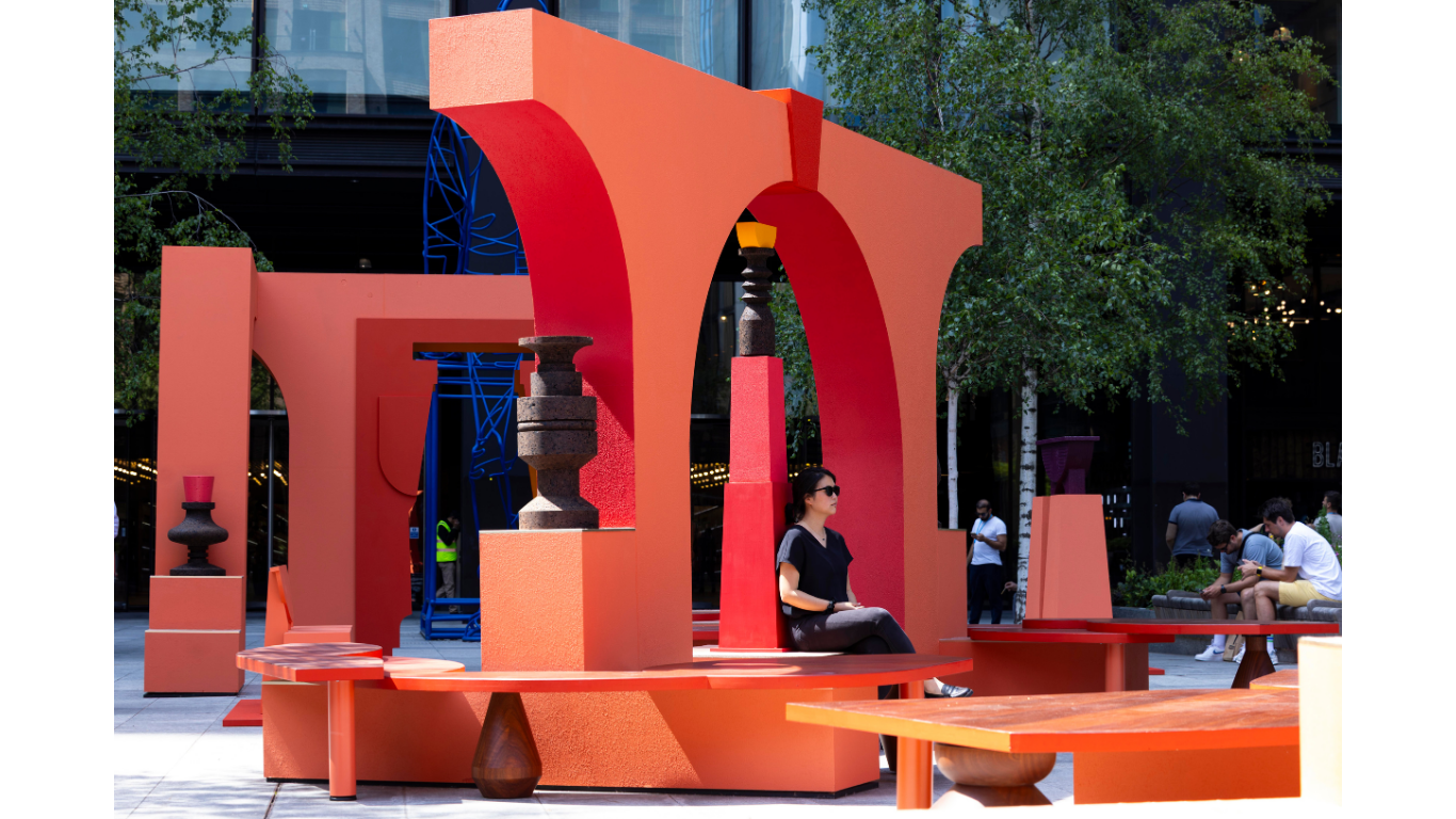 A public art installation in London by Simone Brewster. Architectural forms in shades of red/orange make up a two large structures that resemble temples, the installation boasts archways, seating area and ledges that hold decorative cork sculptures.