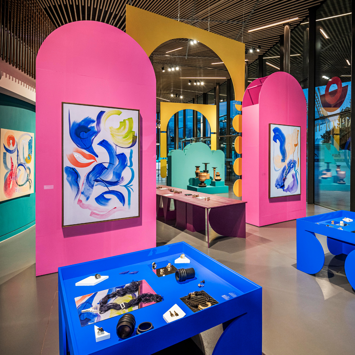 Inside the exhibition paintings hang on two large, bright pink sculptural panels. In the foreground, jewellery is displayed on two Klein blue tables designed by the artist Simone Brewster. A display of the Tropical Noire collection in the background.