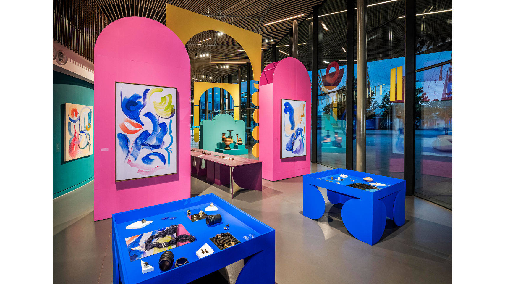 Inside the exhibition paintings hang on two large, bright pink sculptural panels. In the foreground, jewellery is displayed on two Klein blue tables designed by the artist Simone Brewster. A display of the Tropical Noire collection in the background.