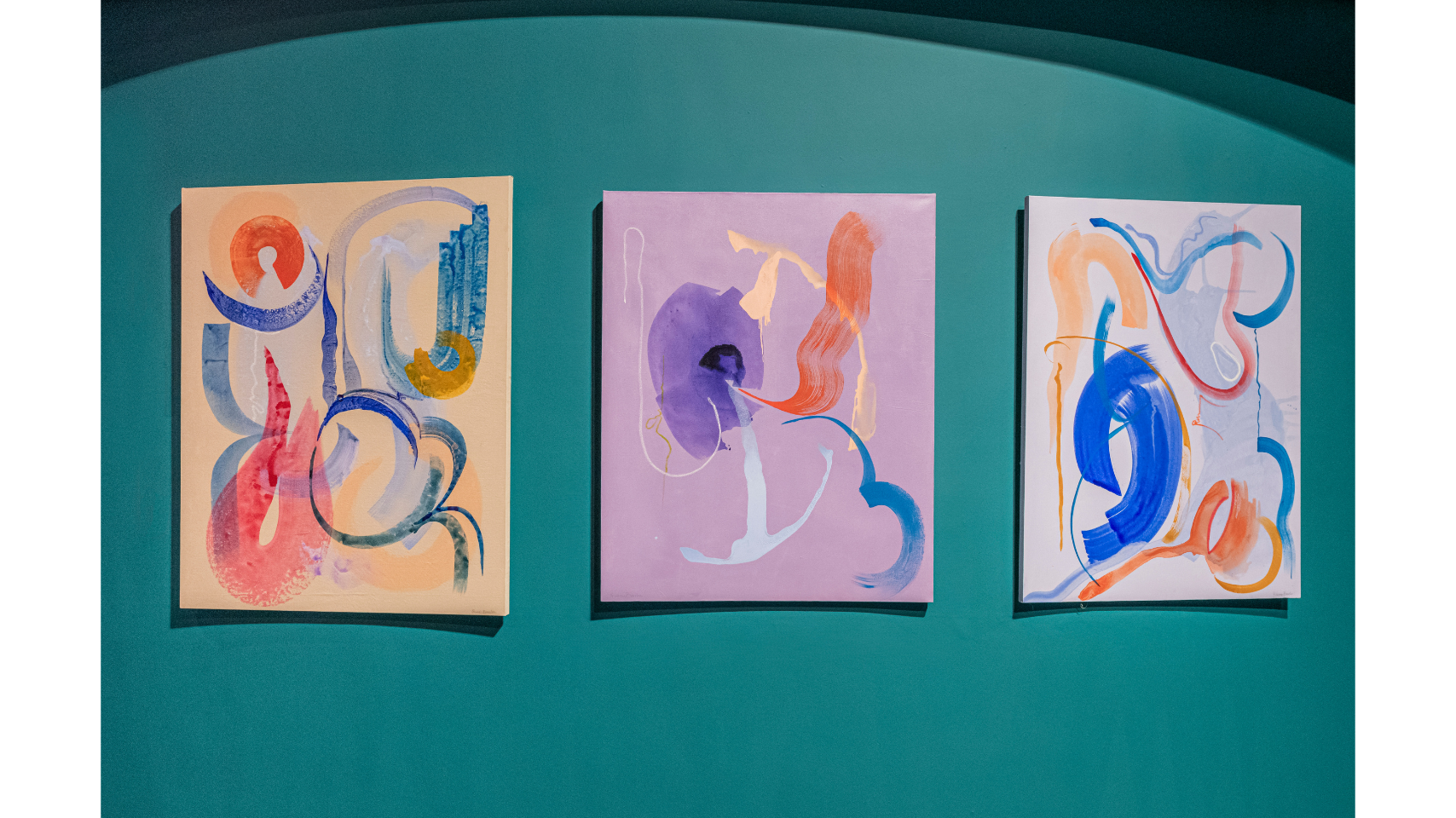 Three colourful paintings are hung up in a row on a turquoise coloured wall.