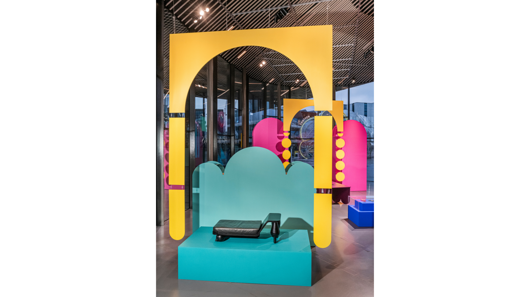 Inside the exhibition space, a chaise lounge designed by Simone Brewster is displayed on a bright, turquoise coloured podium. A large, bright yellow installation in the form of a fragmented arch hangsabove the podium. 