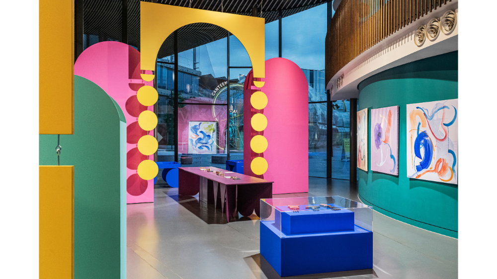 Inside the exhibition colourful paintings hang on turquoise coloured wall. Large colourful installations decorate the space. In the foreground, the Crowns collection is displayed on a Klein blue table covered by a glass case.