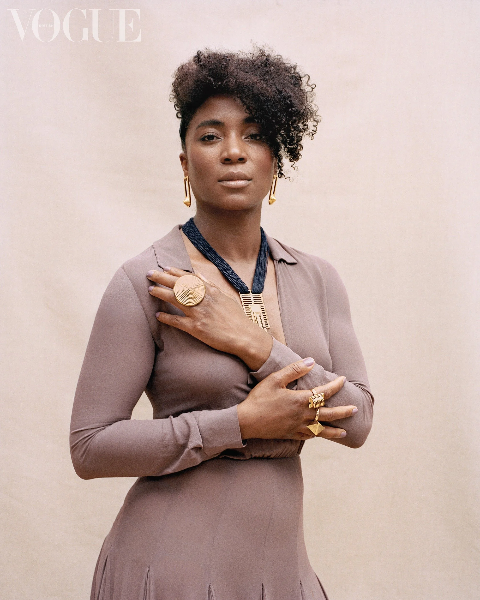 Artist Simone Brewster is wearing gold jewelry, including earrings, a large ring, and a necklace (designed by the artist), poses for a Vogue photoshoot against a light pink background. Feature for Black British Jewellery Design in Vogue.