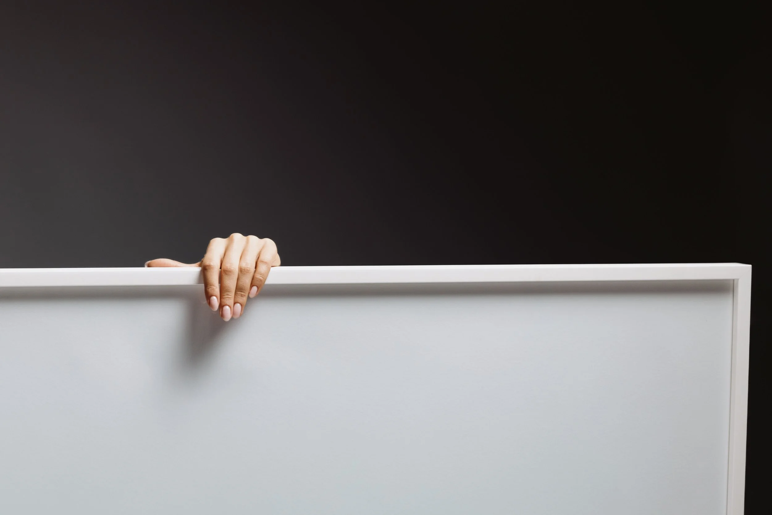 A person's hand peeking over a white cabinet or countertop against a black background.