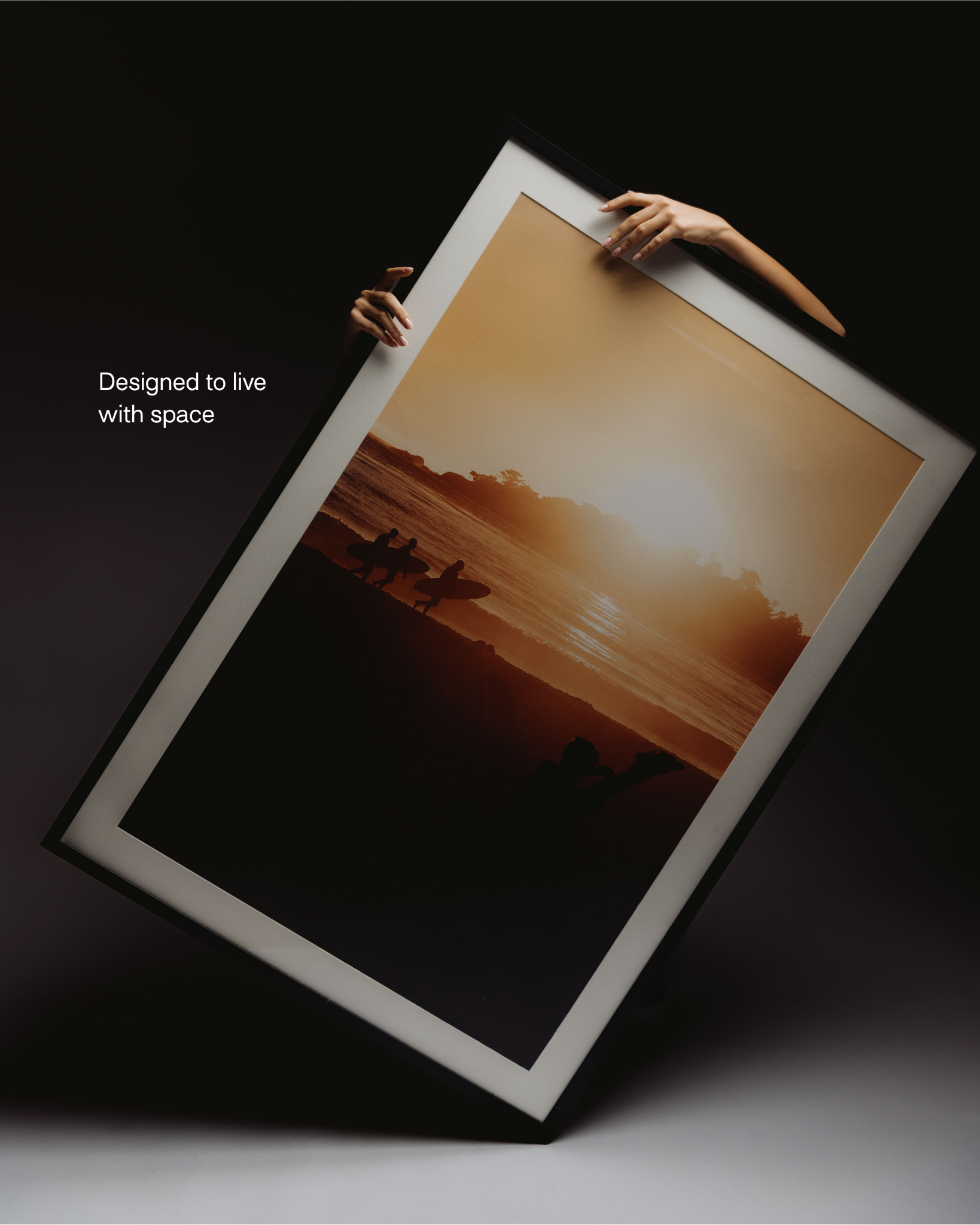 A person holding an illuminated framed sunset photograph of people surfing on a beach at sunset, with the text 'Designed to live with space' on a black background.