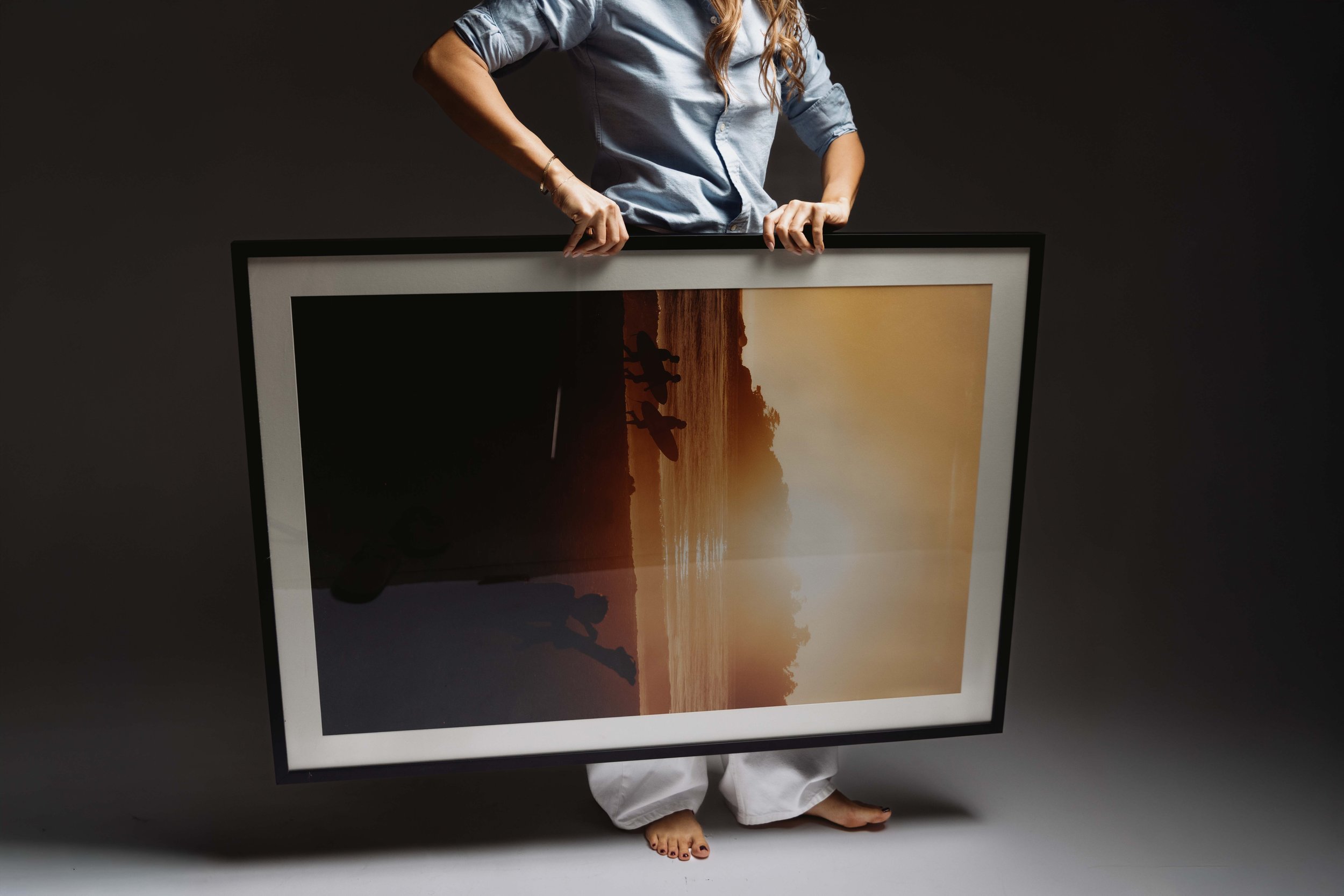 Person holding a large framed photograph of a sunset over a beach with silhouette figures near the water.