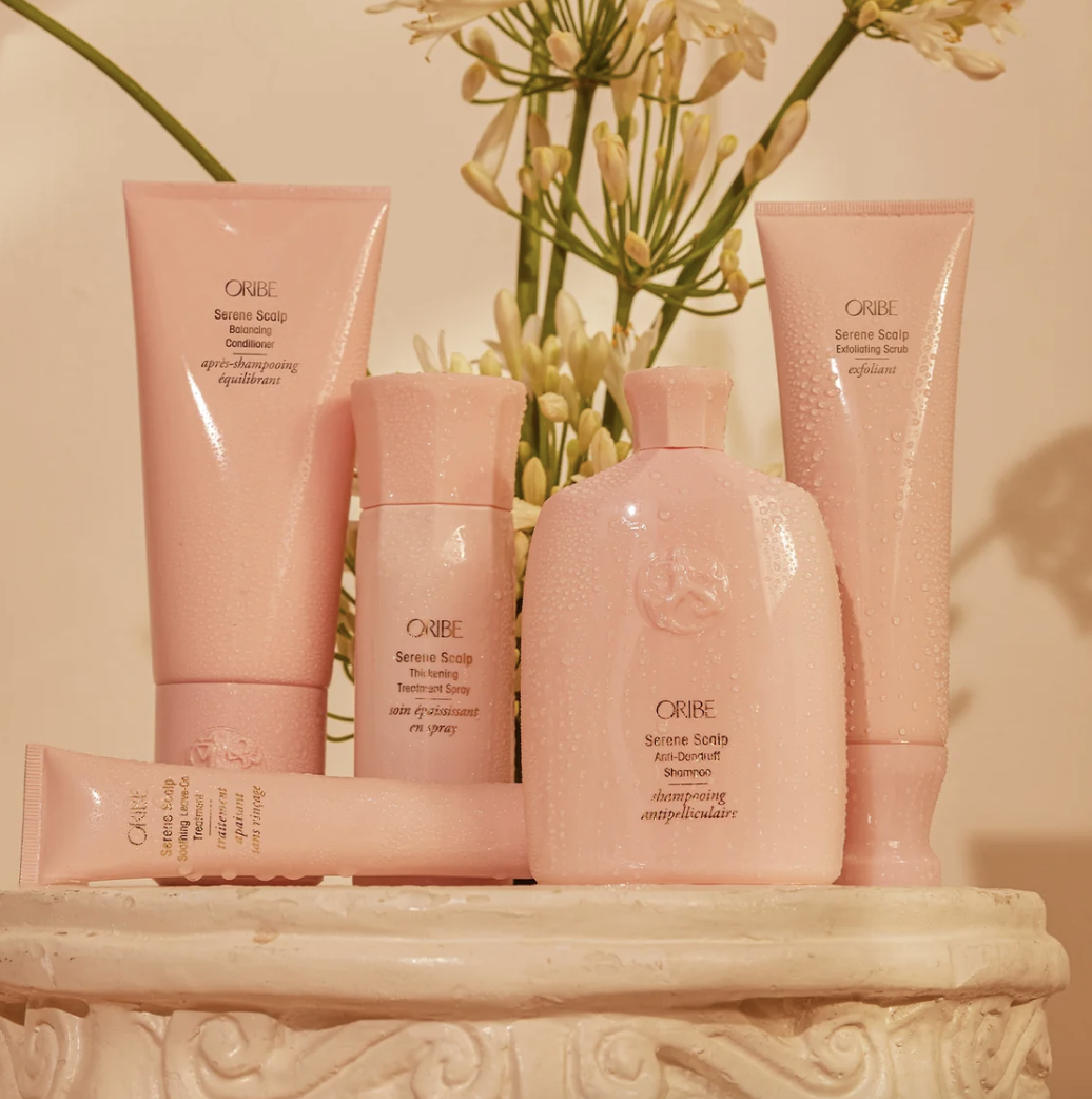 Bild på fem rosa hårvårdsprodukter från Oribe, inklusive schampo, balsam, exfolierande scrub, behandling och spray, placerade på en beiget marmorbord med vita blommor i bakgrunden.