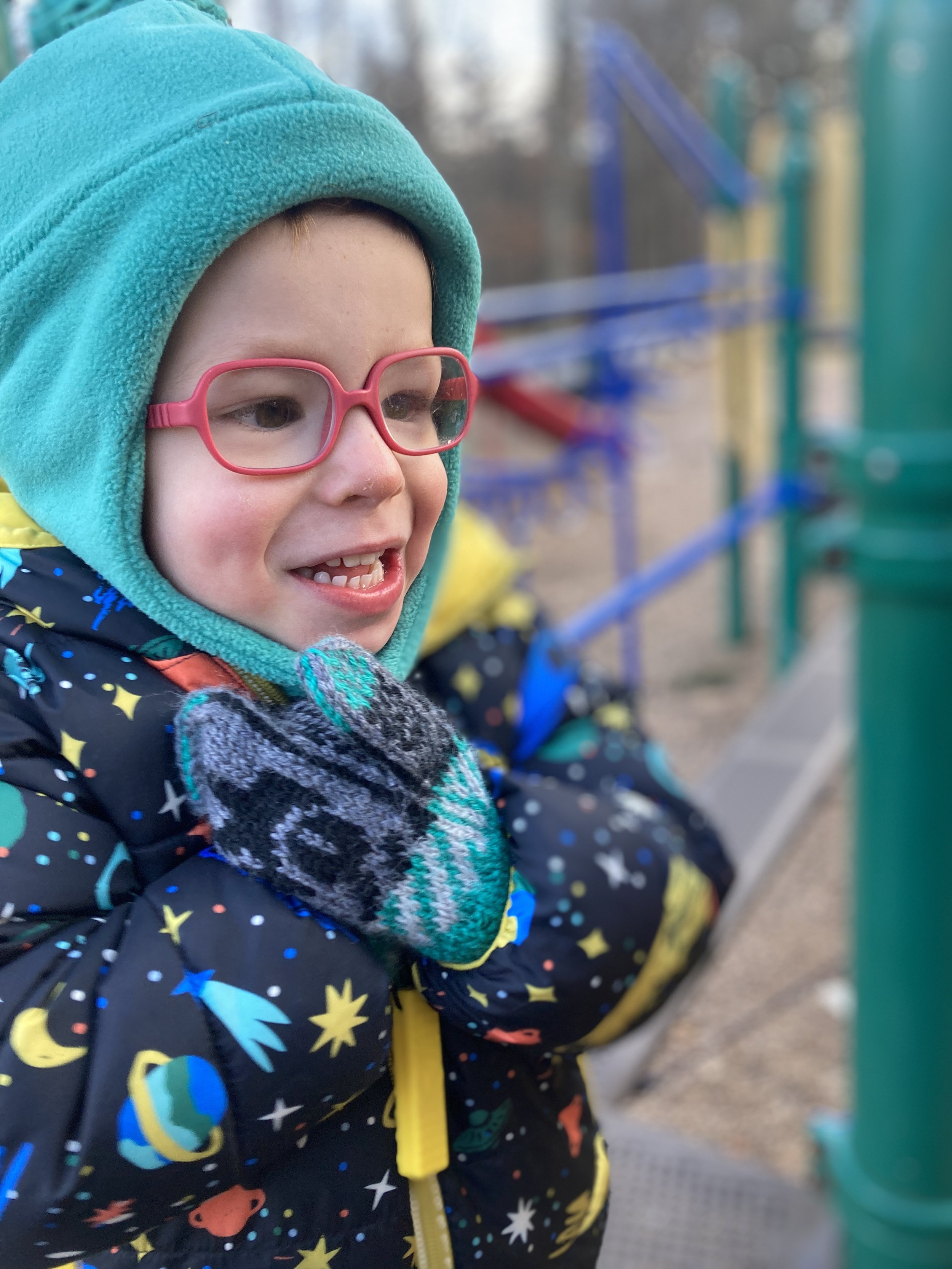 A young child smiling outdoors, wearing pink glasses, a teal hoodie, a space-themed jacket, and gray gloves at a playground.